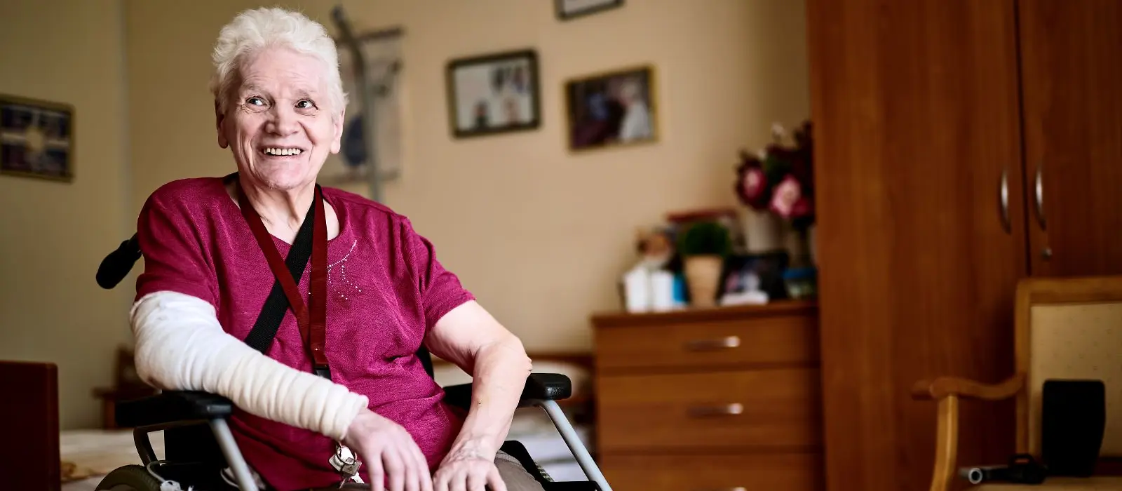 Eine ältere Frau mit einem bandagiertem rechtem Arm sitzt im Rollstuhl und lächelt jemand. an.