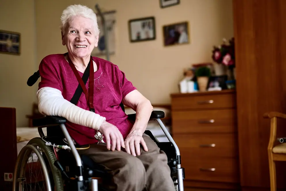 Eine ältere Frau mit einem bandagiertem rechtem Arm sitzt im Rollstuhl und lächelt jemand. an.